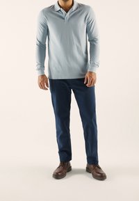 Světle modrá polo košile s dlouhým rukávem a klasickým límcem, spárovaná s námořnickými kalhotami. Model nosí hnědé šněrovací boty.