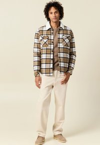 Camicia di flanella a scacchi marrone e bianca con colletto bianco, indossata sopra un maglione beige; abbinata a pantaloni chiari e scarpe beige.
