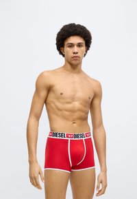 Sarkanas bokseršortus no mīksta auduma ar melnu vidukļa joslu, uz kuras ir uzraksts "DIESEL" un baltas detaļas. Gluda tekstūra un pieguļošs siluets.