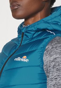 Teal Puffer-Weste mit Kapuze, Reißverschluss und weißen Nähten, mit dem ellesse-Logo auf der Brust, getragen über einem grauen gemusterten Pullover.