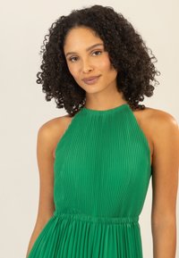 Abito verde a pieghe con scollo all'americana, dotato di un corpetto aderente e una gonna fluttuante, caratterizzato da una vita arricciata e dettagli decorativi sulle spalle.