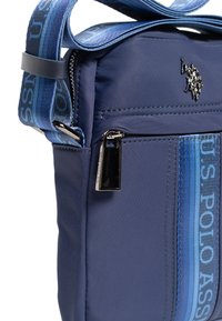 Laivastonsininen nylon olkalaukku, jossa on teksturoitu pinta, hopeanvärinen vetoketju, säädettävä hihna, jossa sininen logo, ja pieni musta tunnus etupuolella.