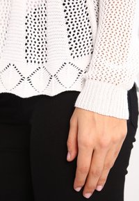 Vit stickad tröja med öppen väv, med diamantmönster vid nederkanten och ribbade ärmslut. Texturen är lätt och andas.