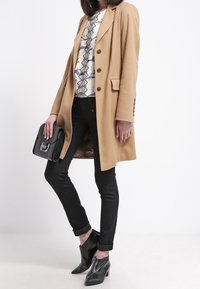 Kamelullkappa med slag och knappar, vit och blå mönstrad blus, svarta tight jeans, svart väska och spetsiga svarta ankelskoot.