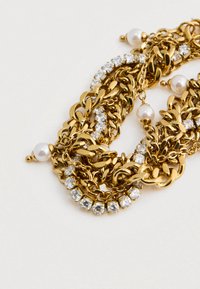 Kultaketju, jonka koristelussa on kirkaita kristalleja ja valkoisia helmikoruja vaalealla taustalla.