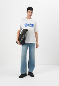 Camiseta blanca con gráfico azul "HugO", combinada con jeans de corte holgado color azul claro y bolso de cuero negro. Botines negros completan el look.