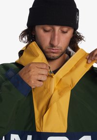 Color-blocked jacket with green, yellow, and navy sections. Features a zip closure, velcro accents, and a prominent logo near the hem.