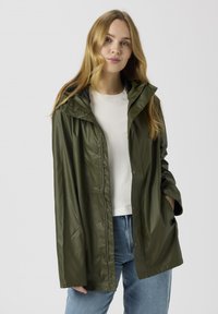 Zöld vízálló cipzáras kabát kapucnival, patentos gombokkal, hosszú ujjakkal és laza szabással. Fehér ing és kék farmer fölött viselve.