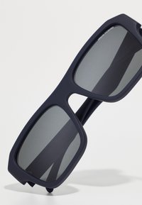 Occhiali da sole oversize blu navy con lenti grigie, finitura opaca e design a cornice geometrica. Hardware minimale e forma angolare.