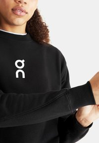 Black sweatshirt with a high neckline and ribbed cuffs, featuring a white logo on the chest. Fabric appears soft with a smooth texture.