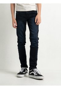 Jeans ajustados de color azul oscuro con un desgaste mínimo, bolsillos frontales y una textura suave, combinados con zapatillas bajas en blanco y negro.