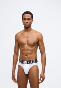 Λευκά βαμβακερά σλιπ με μαύρη ελαστική μέση που φέρει το λογότυπο "CALVIN KLEIN". Λείες υφή και εφαρμοστό σχέδιο. Το μοντέλο έχει γυμνασμένο σώμα.