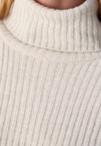Gros plan sur une personne portant un pull à col roulé côtelé de couleur crème avec une texture en tricot douce.