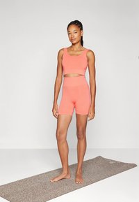 Lengvas, prigludęs sportinis komplektas koralinės spalvos, susidedantis iš trumpu viršutiniu su juosta ir derančių aukštos liemens šortų. Minkšta, lygi tekstūra.