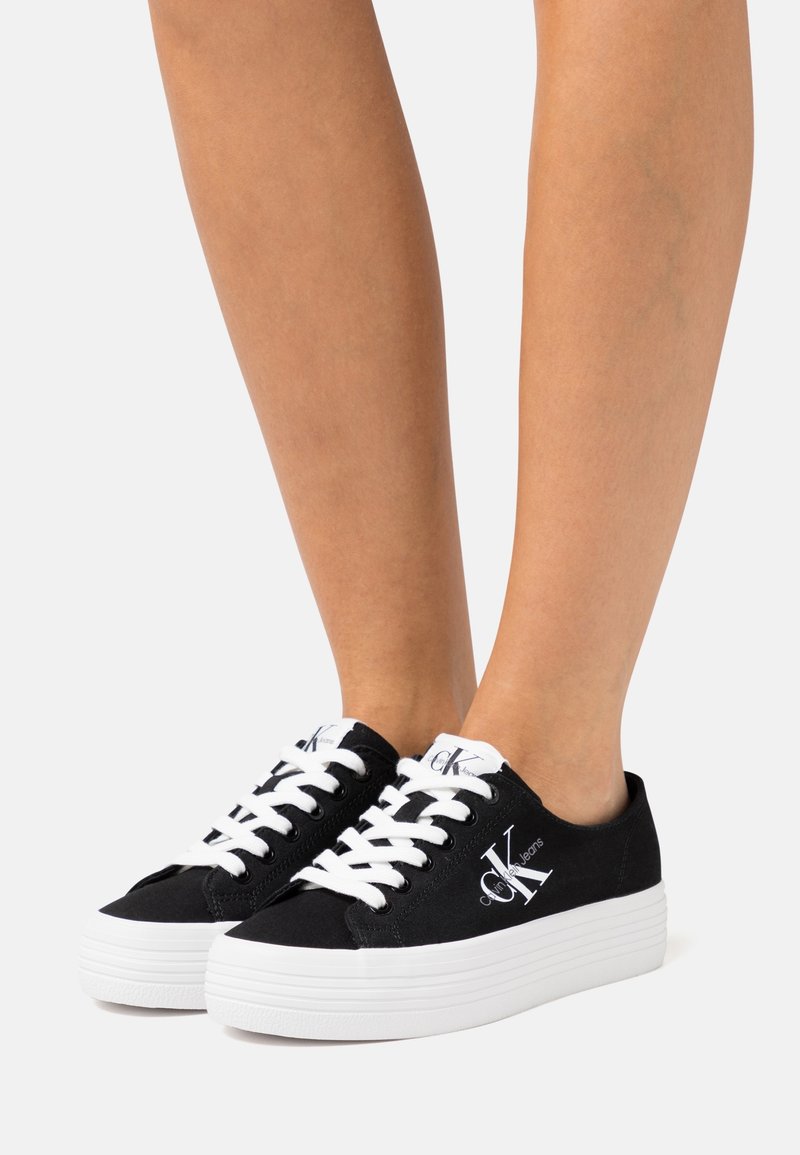 Jalat mengenakan sneakers hitam Calvin Klein dengan tali putih dan sol tebal berwarna putih, di depan latar belakang putih polos.