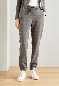 Kvinna iklädd grå skräddarsydd joggingbyxor med elastiska manschetter, guld sidostrecken, vita sneakers och en matchande kavaj, stående på trägolv.