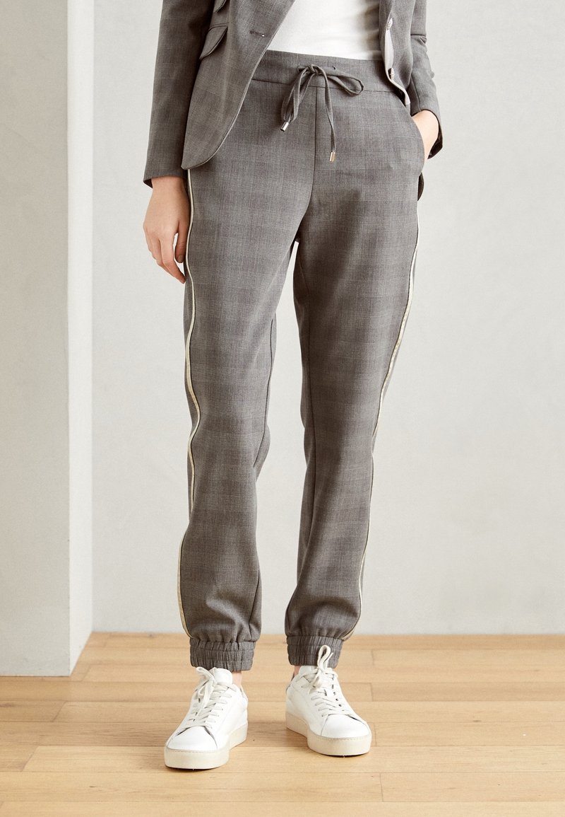 Kvinna iklädd grå skräddarsydd joggingbyxor med elastiska manschetter, guld sidostrecken, vita sneakers och en matchande kavaj, stående på trägolv.