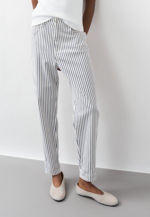 Persona che indossa pantaloni bianchi con strisce nere verticali e scarpe beige senza lacci, in piedi contro uno sfondo bianco e grigio uniforme.