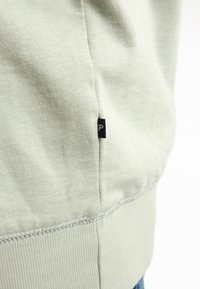 Sudadera verde claro con textura acanalada, dobladillo curvado en la parte inferior y una pequeña etiqueta negra con un logo en la costura lateral.