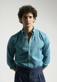 Camisa de botones color teal con botones blancos, mangas largas remangadas, combinada con pantalones de talle alto en color azul marino con pliegues frontales.