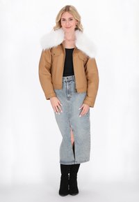 Donna che indossa una giacca beige con colletto di pelliccia bianca, top nero, gonna di jeans a vita alta con spacco frontale e stivali neri, in piedi davanti a uno sfondo bianco.