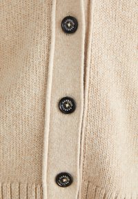 Beige pletený kardigan s texturovanou povrchovou úpravou, který má pět černých knoflíků a žebrovaný lem. Na knoflících je jasné logo značky.