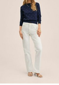 Pull en maille bleu marine avec motif à pois blancs, porté avec un jean blanc taille haute et des talons blancs à lanières. Textures lisses, coupe décontractée.