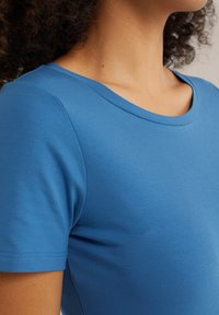 Modré tričko s krátkým rukávem vyrobené z hladkého, elastického materiálu. Má široký výstřih a slim fit, který zdůrazňuje ramena a horní část hrudníku.