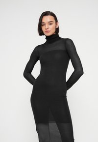 Γυναίκα με κοντά σκούρα μαλλιά, που φοράει εφαρμοστό μαύρο διαφανές φόρεμα ζιβάγκο, στέκεται μπροστά σε απλό λευκό φόντο.