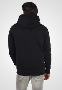 Sudadera negra con capucha, de textura suave y puños acanalados, combinada con jeans de color gris claro, mostrando un ajuste casual desde la parte posterior.