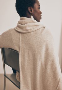 Avvolgente in lana beige con design drappeggiato e morbida texture, dotato di cappuccio e schiena scoperta. Il modello è seduto su una sedia di legno, mostrando la silhouette del capo.