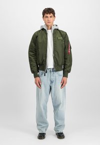Grønn bomberjakke med grå hette, med glidelås, ribbede mansjetter og en sidelomme med detalj av nøkkelring. Brukt over en hvit t-skjorte og jeans.