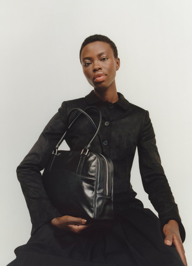 Woman in black buttoned top and skirt holding a large black leather handbag against a plain white background.