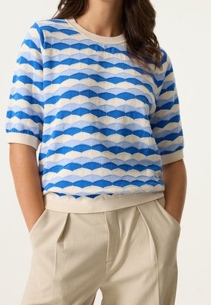 Femme portant un haut en maille à manches courtes bleu et blanc à motifs avec un pantalon plissé beige clair, mains dans les poches, sur un fond uni.