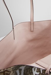 Sac fourre-tout beige ouvert avec des bretelles fines et un tissu texturé à l'intérieur, laissant entrevoir partiellement l'extérieur à motifs avec un grand design textuel.