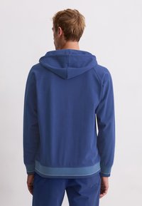 Mann, der wegschaut und einen blauen Kapuzenpullover mit gerippten Bündchen und Bund trägt, kombiniert mit passenden blauen Hosen vor einem schlichten Hintergrund.