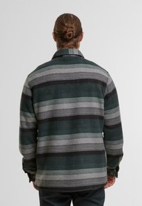 Uomo con capelli castani raccolti in uno chignon che indossa una giacca a maniche lunghe a righe verdi, nere e grigie, mostrato di spalle su uno sfondo semplice.