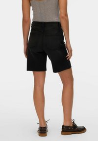 Pantalones cortos de mezclilla negra con un dobladillo sin acabados, que presentan una cintura alta y bolsillos traseros, combinados con zapatos marrones de cordones. El material parece resistente.