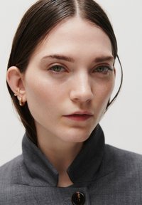Jeune femme aux cheveux bruns lissés en arrière, portant des boucles d’oreilles en or en forme de créoles et une veste sombre à col montant, regardant droit devant avec une expression neutre.