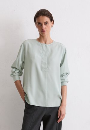 Hellgrüne Bluse mit langen Ärmeln und rundem Ausschnitt, zentralem Knopfleisten-Detail, leicht lockerer Passform und glatter Textur. Kombiniert mit dunklen Hosen.