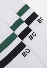 Weiße Socken mit grünen und marineblauen Streifen, die eine gerippte Struktur aufweisen und die Buchstaben "BO" in Schwarz nahe der Oberseite gestickt sind.
