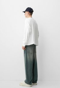 Bela srajca z dolgimi rokavi, sproščenega kroja, v kombinaciji s širokimi, zbledelimi jeans hlačami. Oblečena z temno modrim klobukom in off-white supergami.