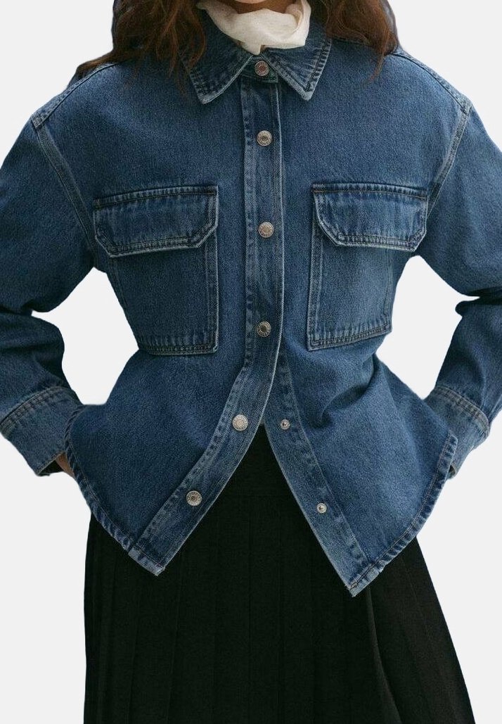 Femme portant une veste en denim bleu boutonnée avec deux poches poitrine et une jupe noire plissée, les mains posées sur les hanches.