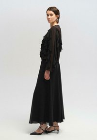 Juoda ilga suknelė su sluoksniuotomis volanų rankovėmis, plaukiojanti siluetas, pagaminta iš skaidraus audinio, derinama su juodais smailėjančiais kulnais.