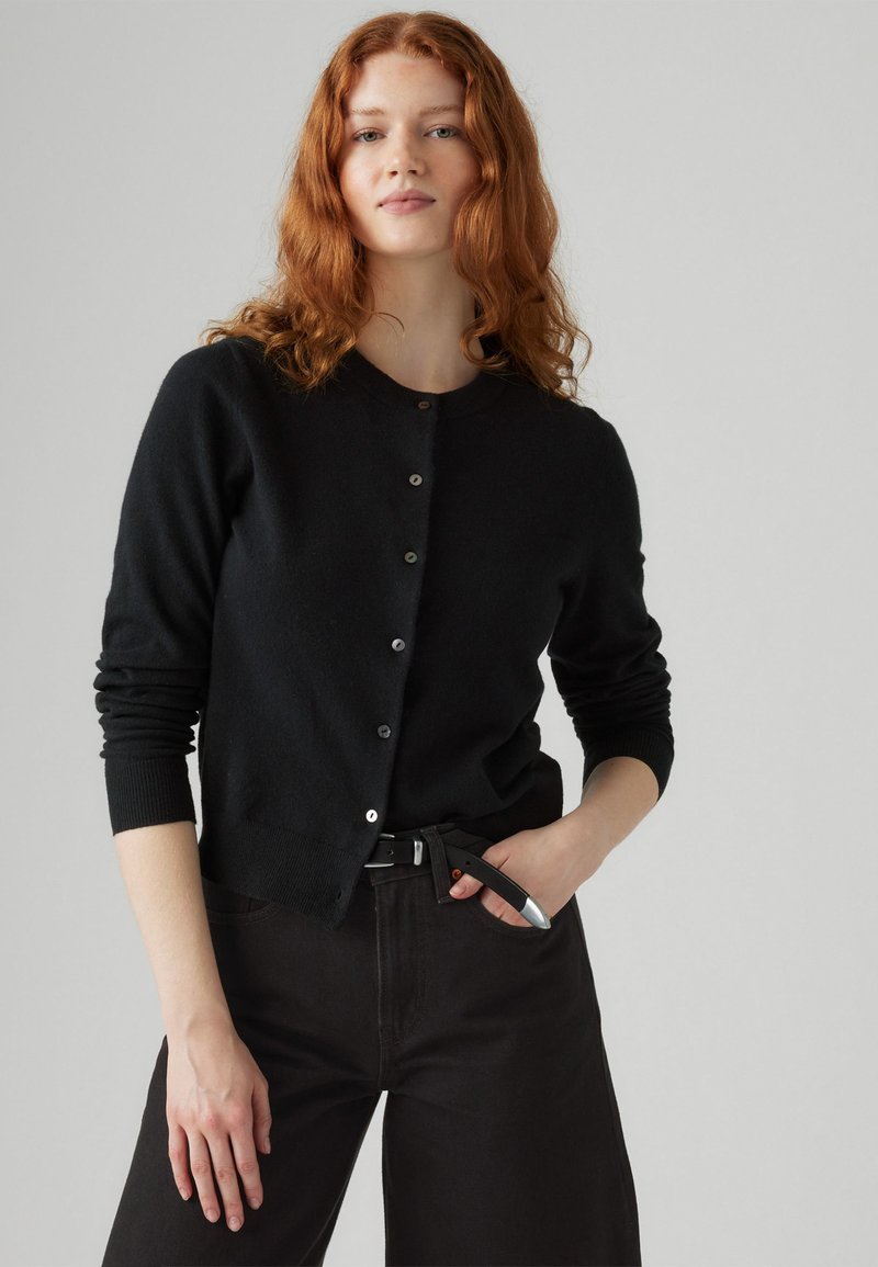 Zwarte gebreide cardigan met een ronde halslijn, lange mouwen en knoopsluiting. Gecombineerd met zwarte high-waisted broek en een riem.