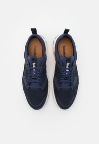 Ett par mörkblå Timberland-sneakers med vita sulor, marinblå snörning och tan innersulor, sedda ovanifrån på en vit bakgrund.