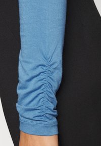 Blå långärmad topp med rynkad textur vid underarmen, kombinerad med slät svart tyg på sidan. Smoothing, lätt material.