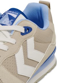 Beige und weiße Sportschuhe mit strukturiertem Obermaterial, blauen Akzenten und einem gepolsterten Schaft. Merkmale sind Perforationen und kontrastierende Logo-Details.