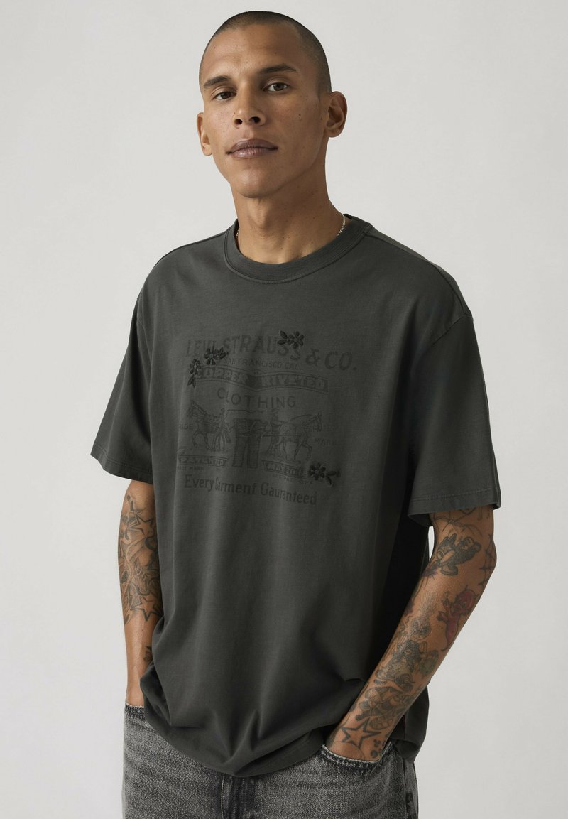 Homme aux cheveux courts et tatouages, portant un t-shirt graphique noir oversize et un jean gris, debout les mains dans les poches devant un fond uni.