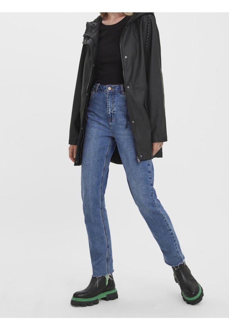 Giacca nera impermeabile con cappuccio, abbinata a jeans blu a vita alta e stivali neri robusti con suole verdi. Design semplice e casual.